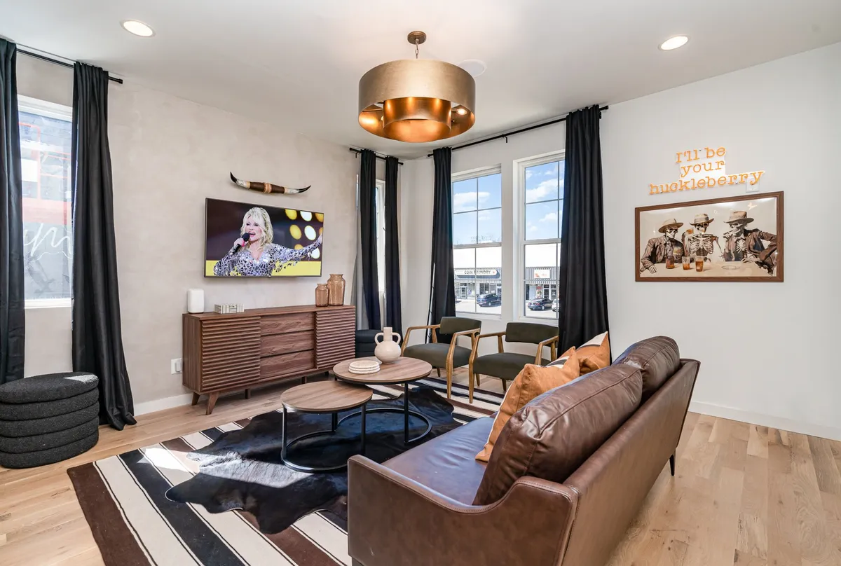 The image showcases a stylish living area featuring a plush leather sofa, a modern coffee table, vibrant Southwestern-inspired artwork, and a large TV displaying a lively performance. The space is designed for comfort and togetherness, perfect for relaxing or socializing.