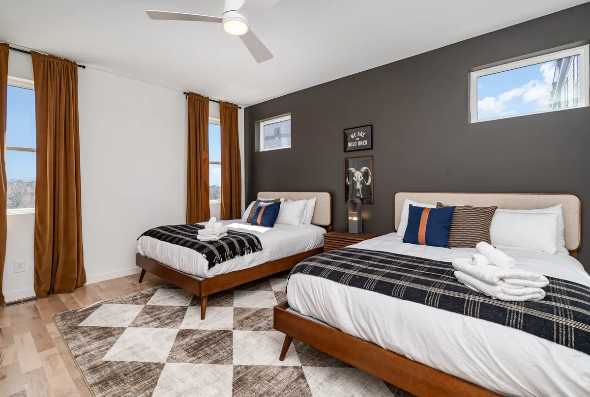 This image shows a bedroom with two beds adorned with decorative pillows, stylish wall art, and large windows that allow for plenty of natural light. The modern ceiling fan and cozy linens add a welcoming touch, while the overall decor feels both modern and inviting.