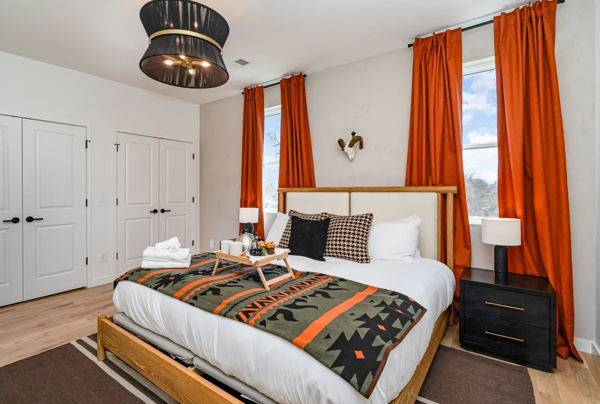 A bedroom featuring a bed with a stylish patterned blanket, bold orange curtains, a modern light fixture, and decorative wall art. The cozy ambiance is enhanced by the soft lighting and attention to detail in the decor.