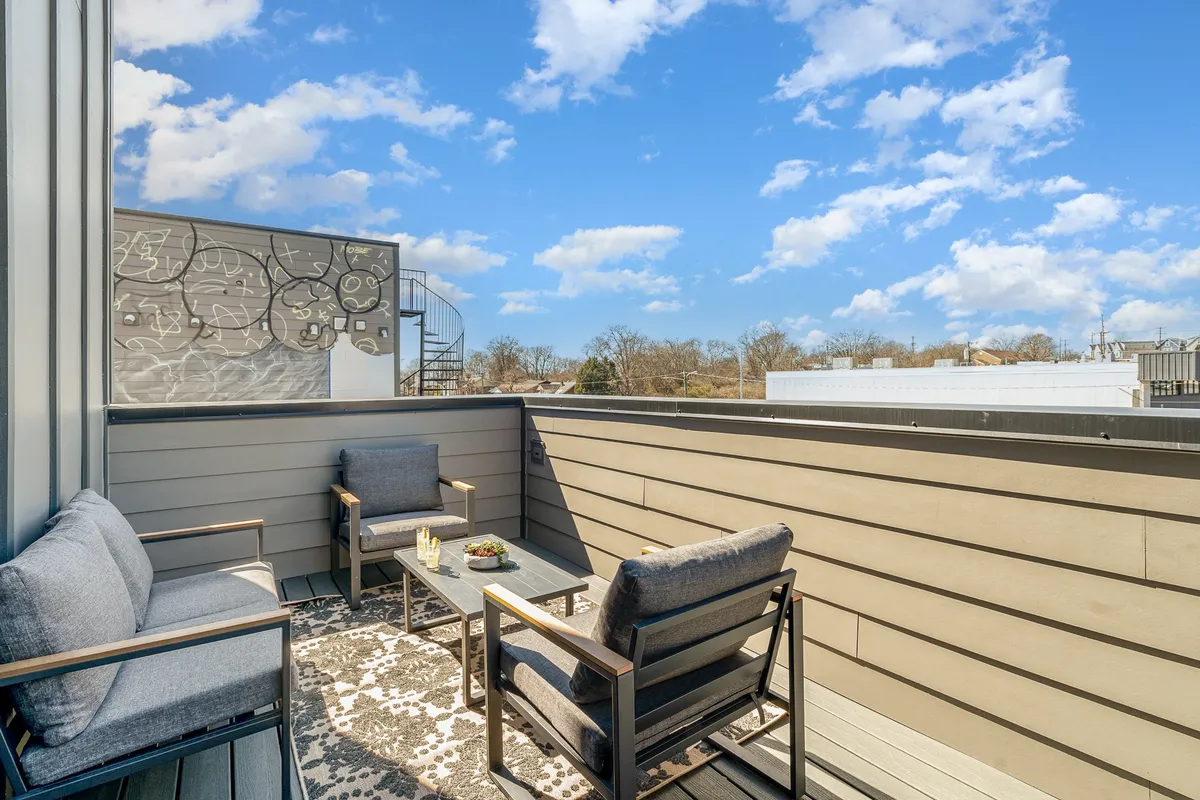 A cozy rooftop deck featuring a decorative rug, a spiral staircase leading up, an inviting outdoor seating area, and the urban Nashville skyline visible in the background. The setting is optimized for relaxing, socializing, and enjoying meals while taking in panoramic city views.